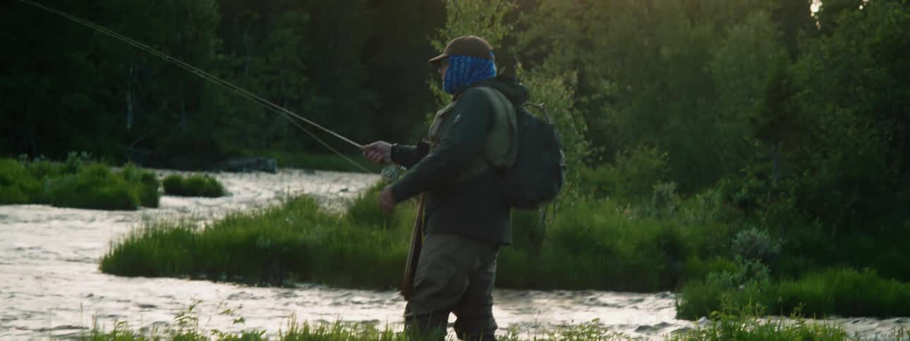 A man with a hat and backpack fishing in the river with a fishing rod with trees the sky and clouds in the background