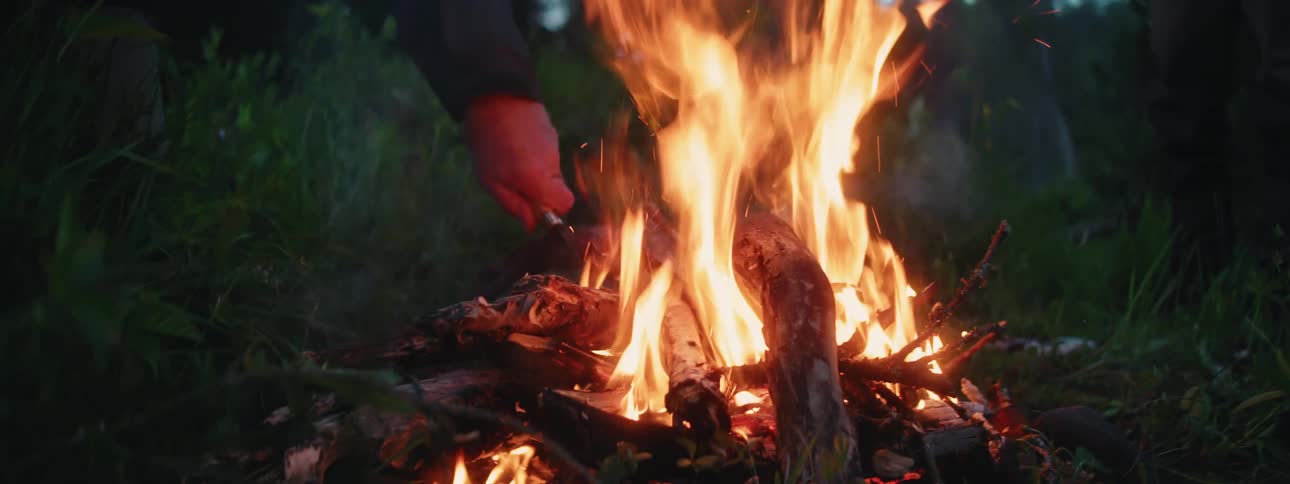 A man with a hat and glasses stoking a fire with a knife with trees the sky and clouds in the background