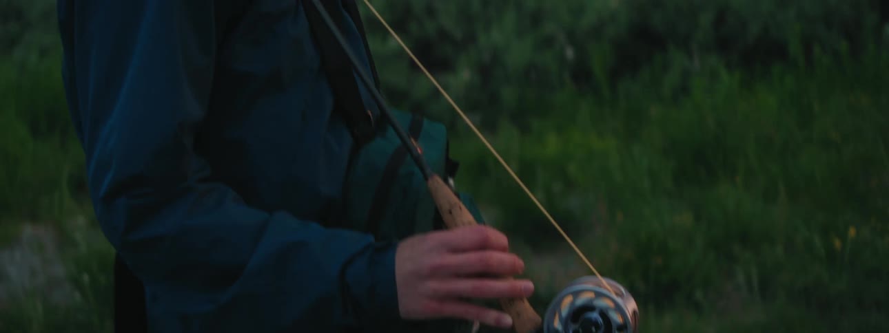 Young man carrying a fishing rod standing near a river in the forest at sunset