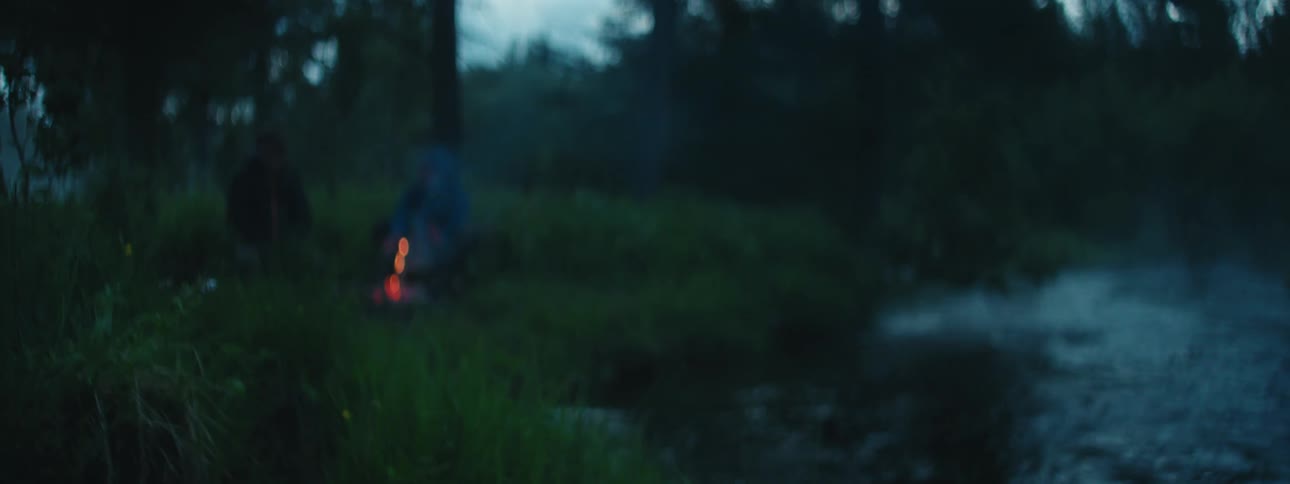 Two men keep warm over a fire while camping in the woods near a lake