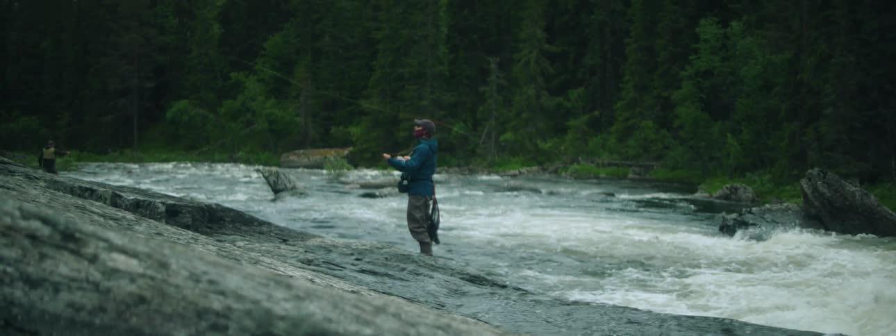 A man walks down the rocky riverbank slope into the flowing water and casts his fishing line