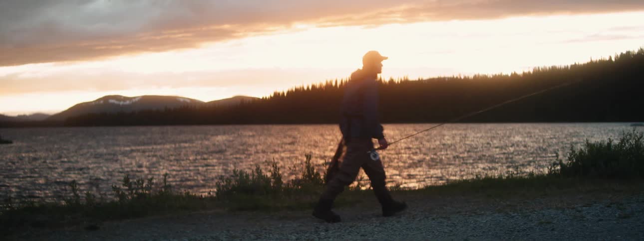 A fisherman carries his fishing pole and net while walking along a riverbank