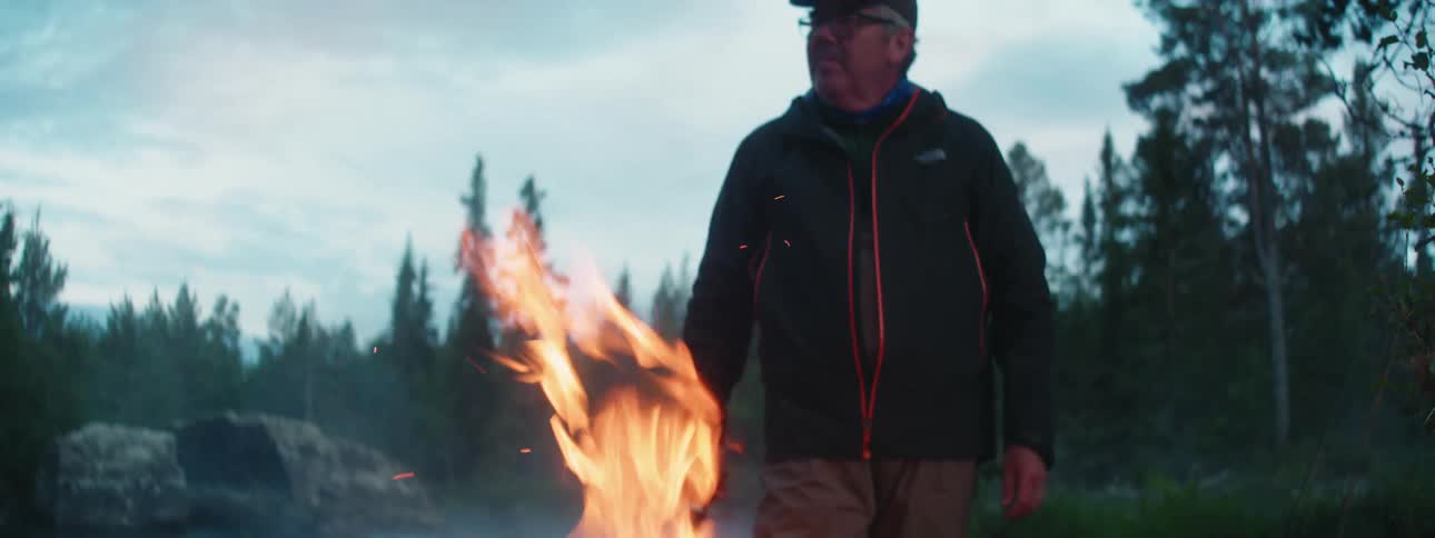 A man uses a long knife to rearrange branches in a campfire beside a river in the wilderness on a cloudy day