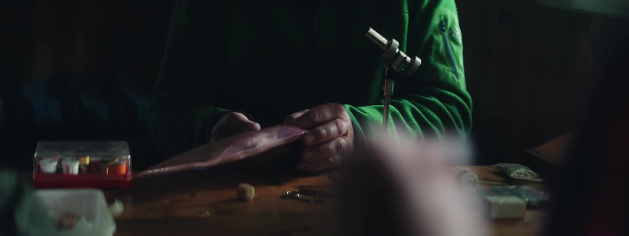 A man and two others are sitting indoors at a table producing handcrafts during the day