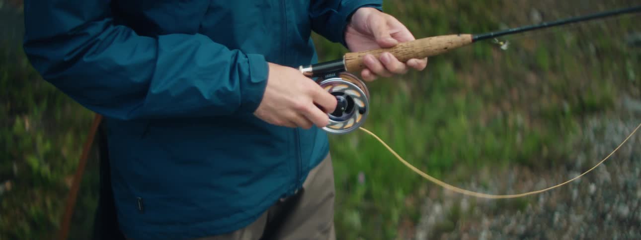 A fisherman slowly winds up the fishing line of his rod while he fishes at a river