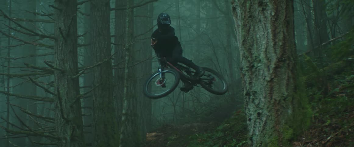 A mountain bike athlete whips their bike sideways off a foggy forest jump