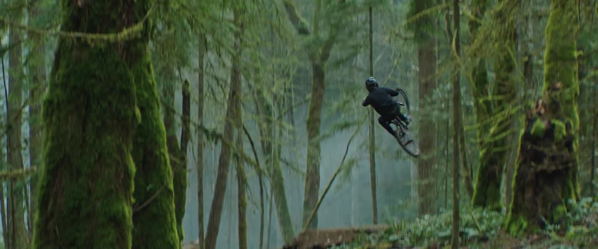 A mountain biker hits a large wooden ramp in the forest and whips his bike sideways in the air
