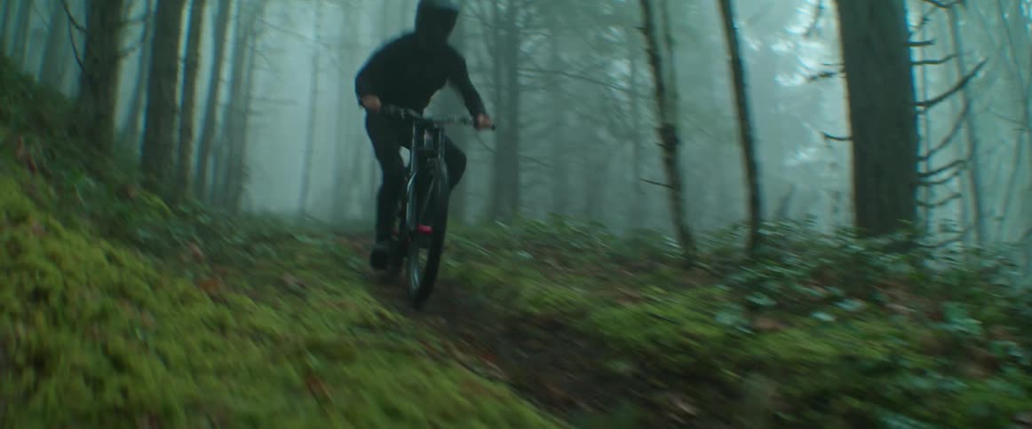 A cyclist rides their mountain bike on a foggy forest trail, kicking up dirt towards lens