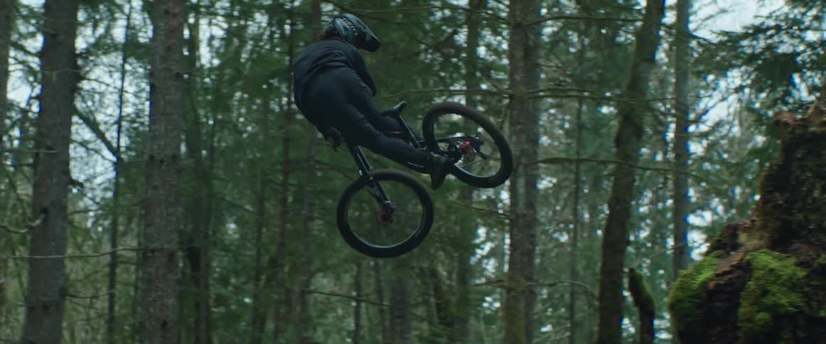 A man whipping his mountain bike through the air off of a large wooden jump in the forest