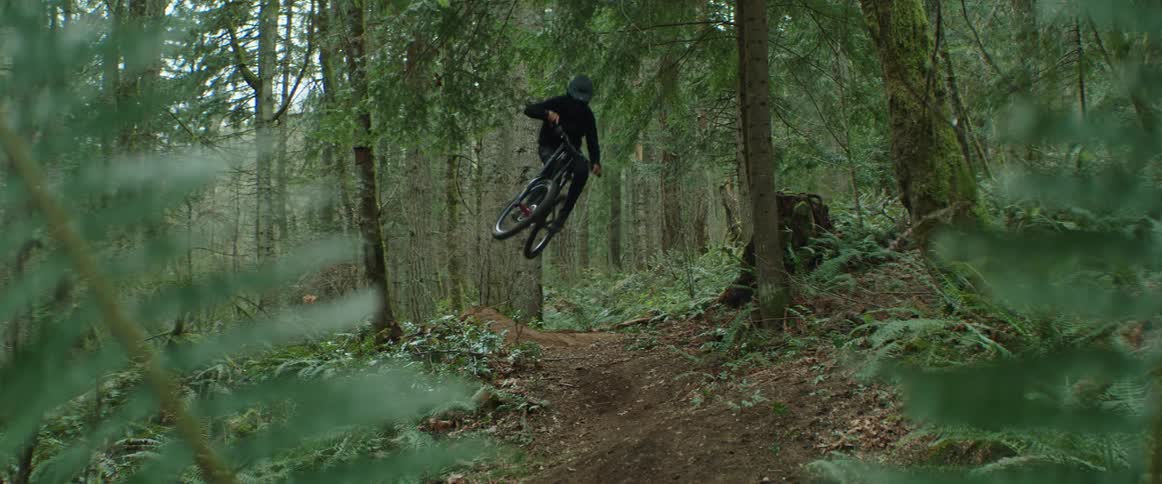 A cyclist goes airborne while riding at high speed along a dirt trail through a forest during the day