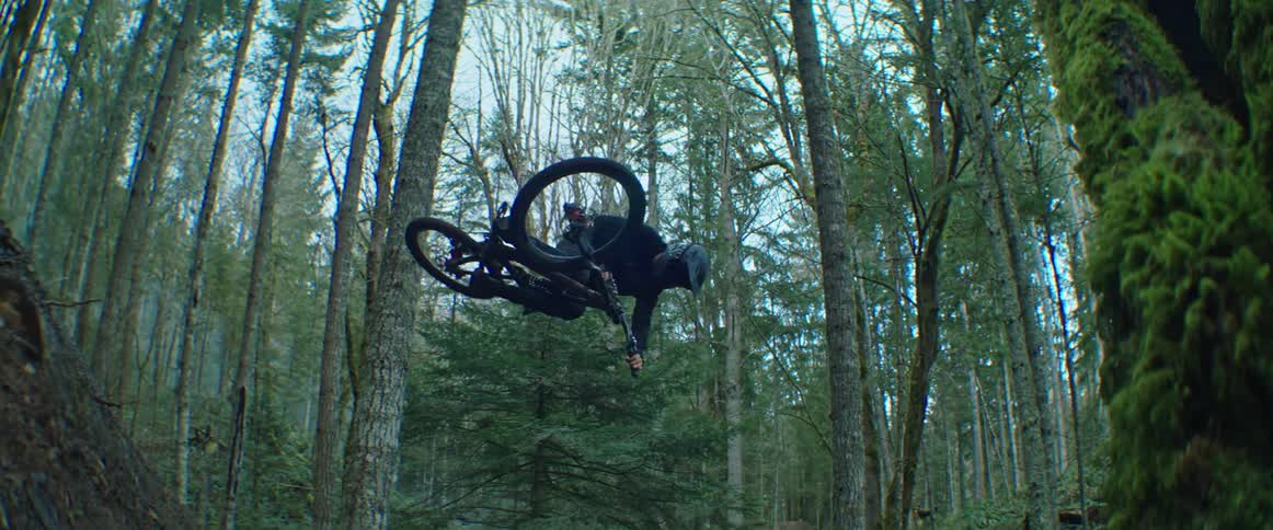 A person does a large jump on his mountain bike and performs a tabletop trick in the forest