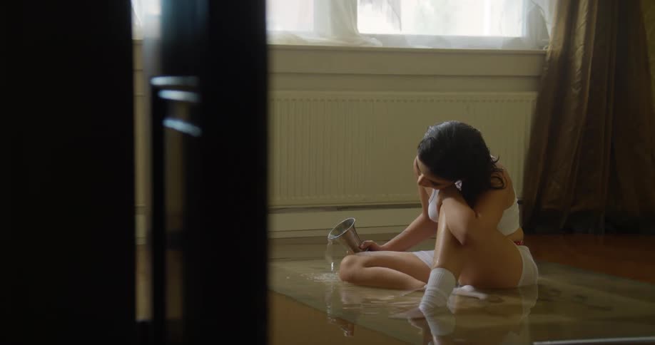 A young woman sitting on the floor of a flooded room while holding an overflowing cup