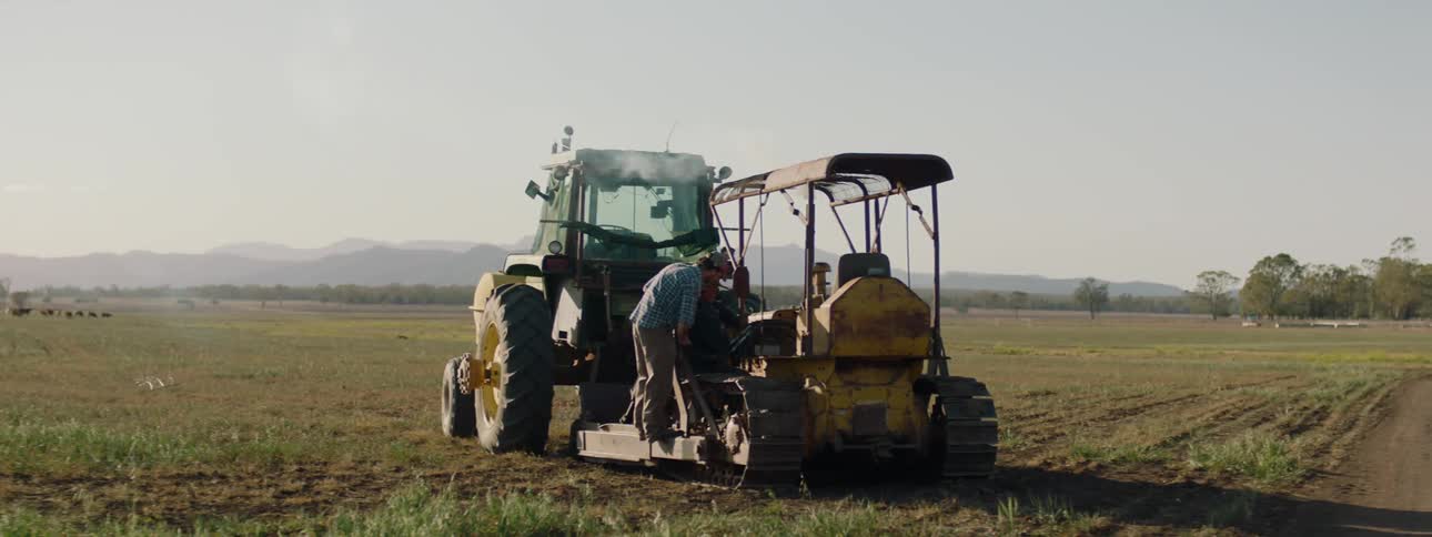 Two men stand talking over smoking vehicle attached to tractor with cattle and foggy mountains in background