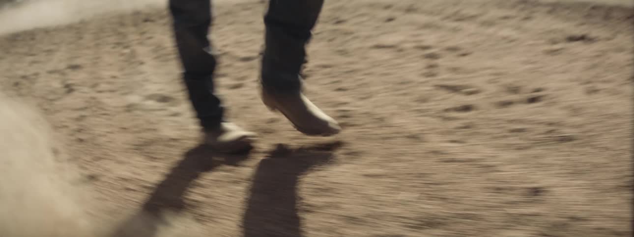 A man in cowboy boots kicks up dirt as he walks through a field