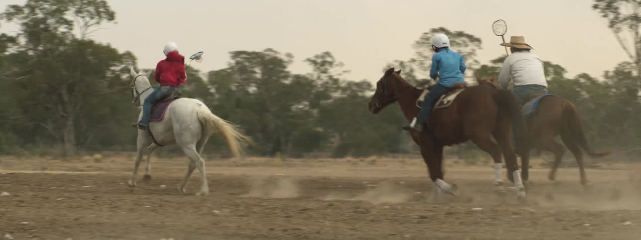 Riders play game with netted hoops and a ball riding their horses across dirt field as dust blows by
