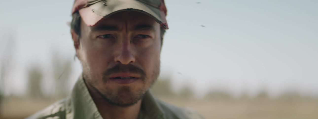 A man with a hat with flies swarming all over his face with the sky and clouds in the background