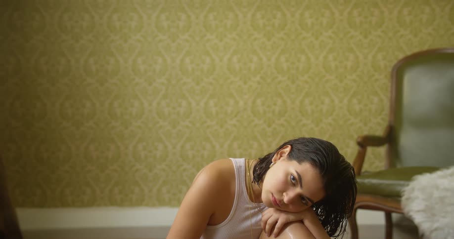 A young woman sitting on the floor with her head resting on her knee glances up into the camera