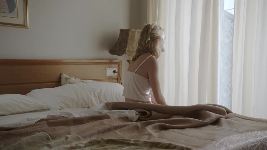 A woman sits on the edge of a bed looking out a window