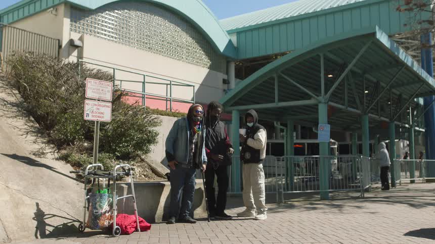 A man leans heavily on his cane standing with others outside of a bus station