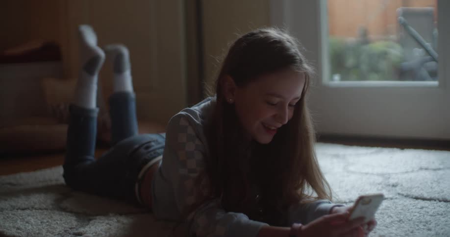 A girl smiles while talking on a video call lying on a rug kicking her legs