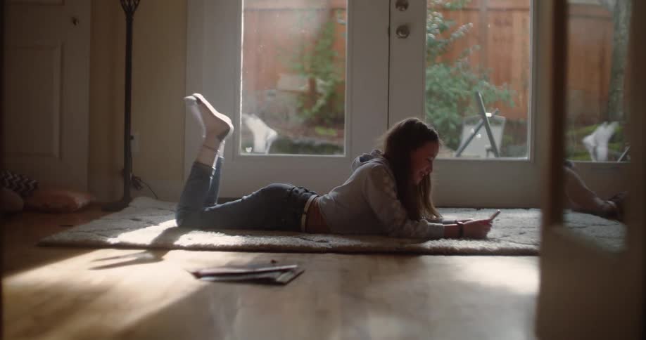 A girl lying on her stomach by the french doors waves to her phone as she kicks her legs