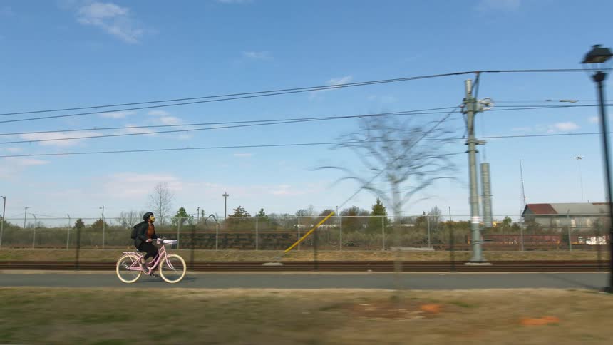 Woman pedals along street on bicycle passing street lamps and a yellow fire hydrant with building in fenced area in background