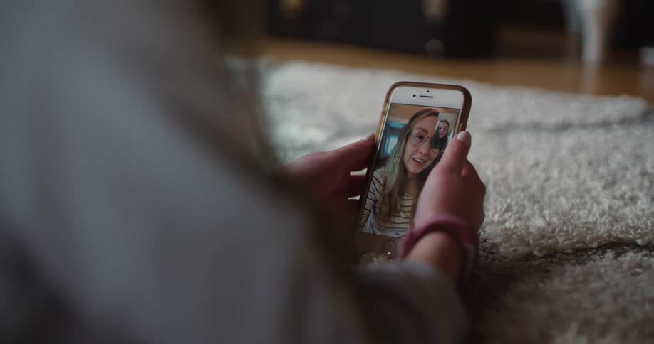 A girl lies on a rug holding a smartphone having a facetime call with a friend