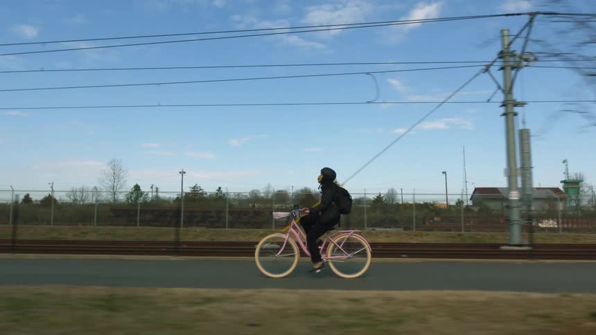 Woman wearing helmet and backpack pedals bicycle along street past fenced grassy field