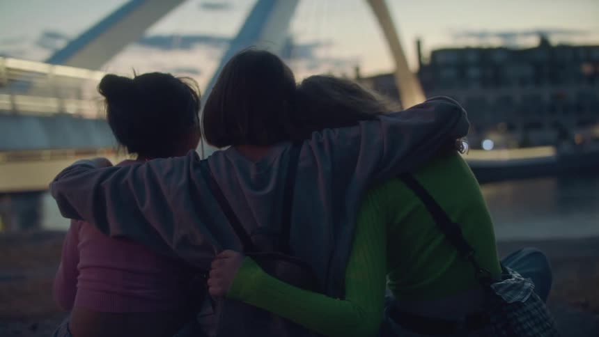 Three teenage girls hugging outside near a city bridge at sunset