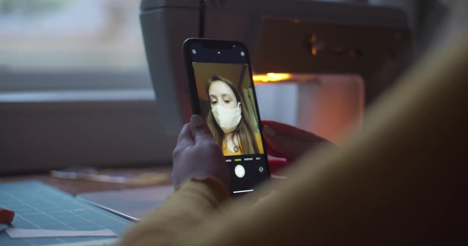 A young woman sitting at a desk indoors uses her smartphone to take a photograph of her masked face