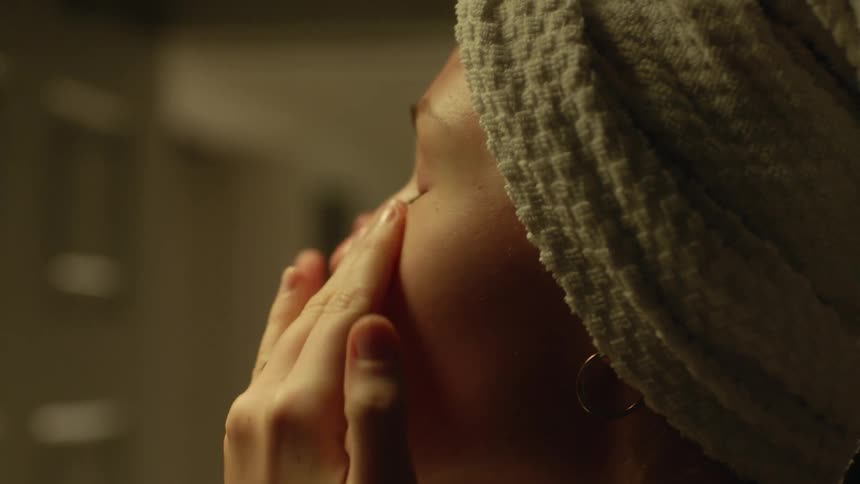A woman with hair wrapped in a towel smooths her fingers over her eyes and brow