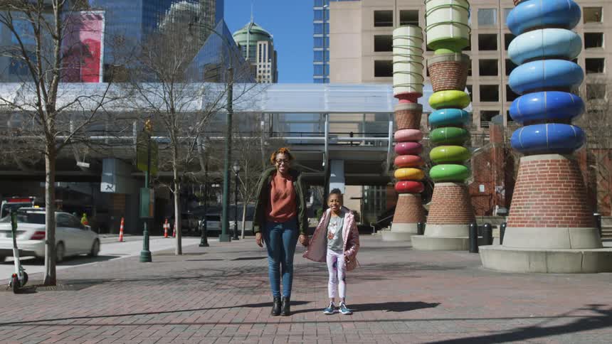 A mother holding her young daughters hand on a city sidewalk with skyscrapers in the distance