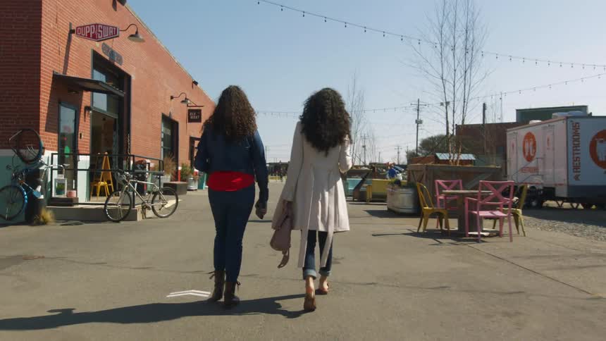 Two women walk together outdoors near a building with an outdoor dining table nearby