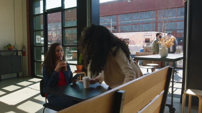 A woman walks around a restaurant and sits down at a table with a friend
