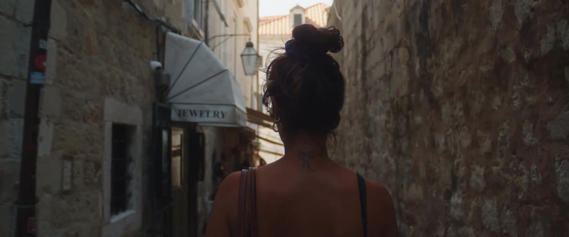 A woman with a tattoo on the back of her neck walks down a narrow alley past stores on a bright sunny day