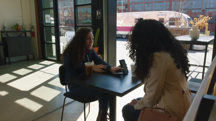 A woman shows her phone to her friend in a cafe