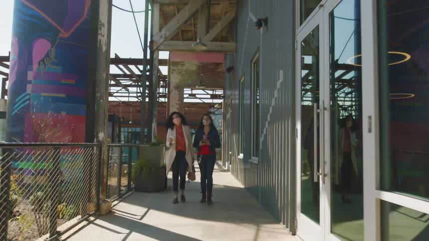 Two women carrying coffee cups walk past a painted creative building on a sunny day