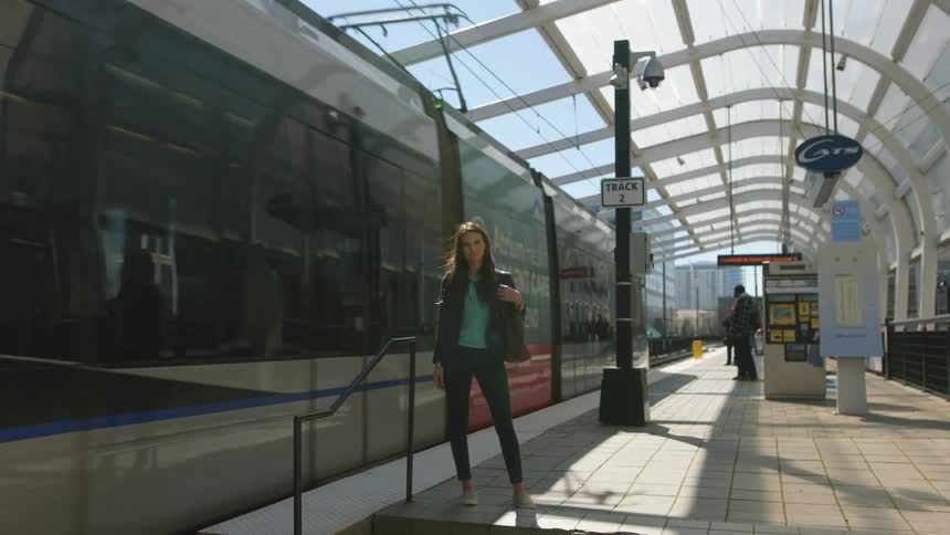 A young woman is standing near the tracks in a train station as a train passes by her
