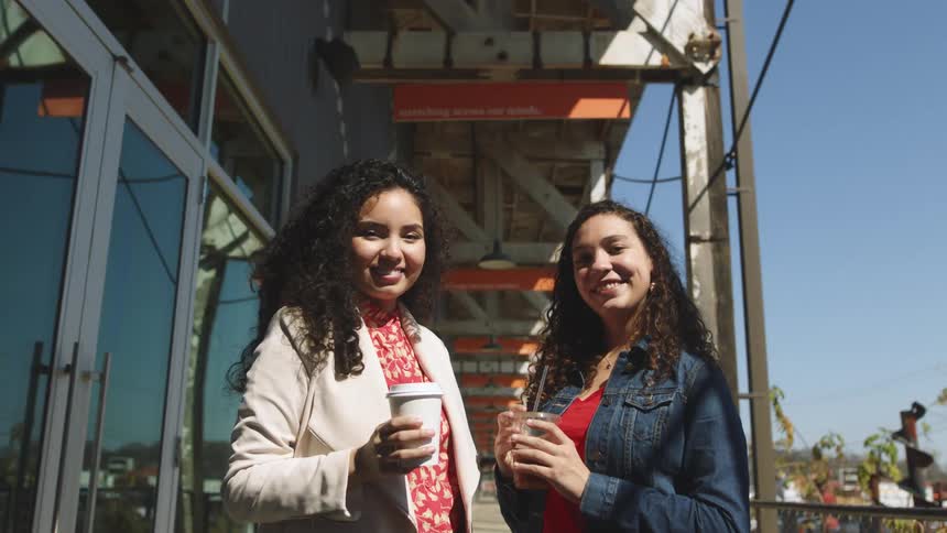 Two women standing and holding cups next to a building with the sky and clouds in the background