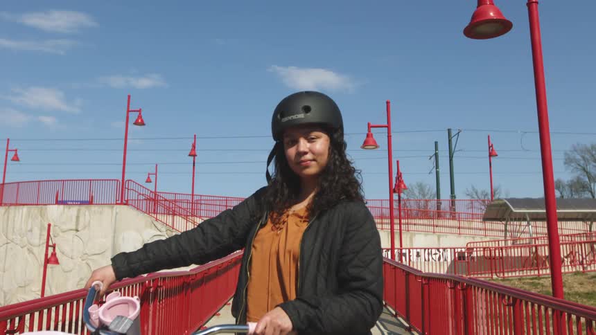 Girl wearing a black helmet and jacket is on a bicycle with a red fence and red lamp posts around the area