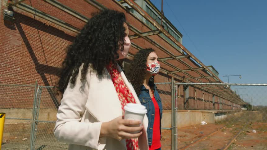 Two women walking with mask with a fence a building the sky and clouds in the background