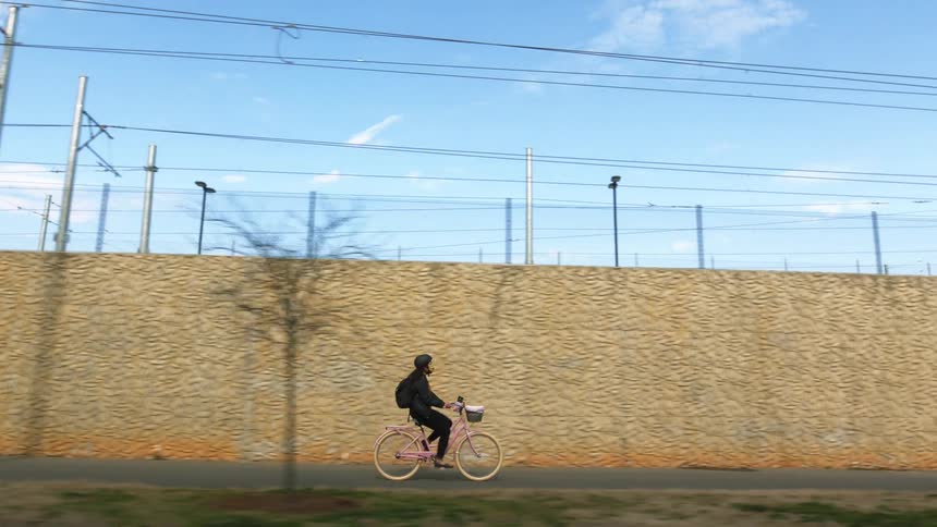 A person with a helmet and a backpack riding a bicycle with a wall the sky and clouds in the background