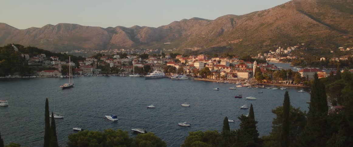 Boats are anchored off the coast in a harbor near a village with hills behind