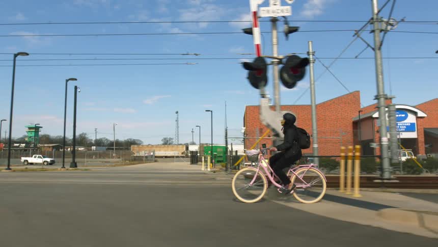 A person wearing a mask pedals a bike past a railway crossing on a bright sunny day