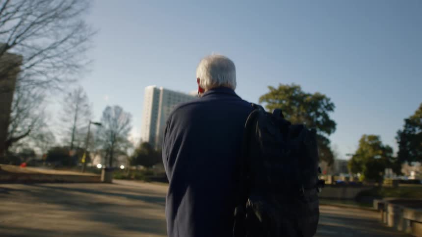 Older white haired man carrying backpack walks across plaza in city