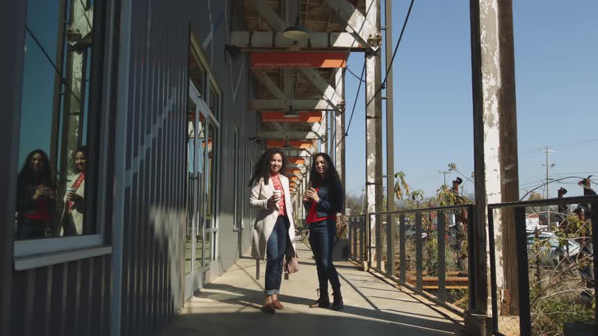 Two women holding portable drink cup stand near building under covered walkway smiling as car passes in background