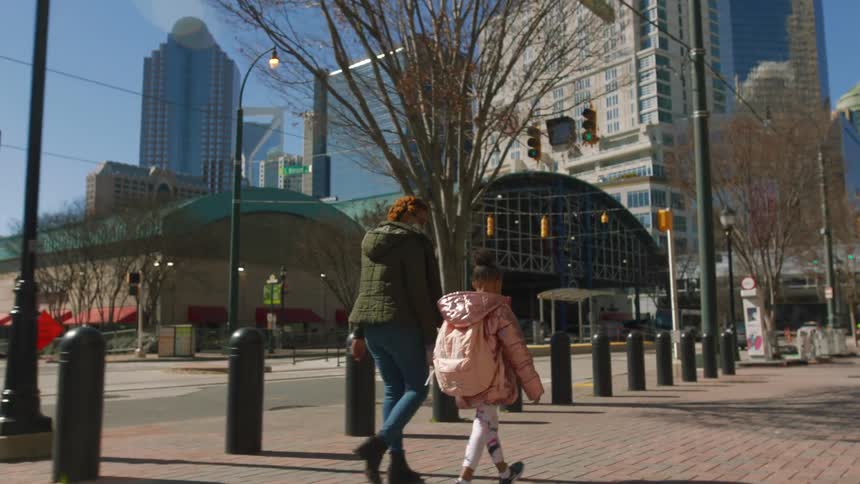 A mother walks with her daughter on a city sidewalk on a bright sunny day