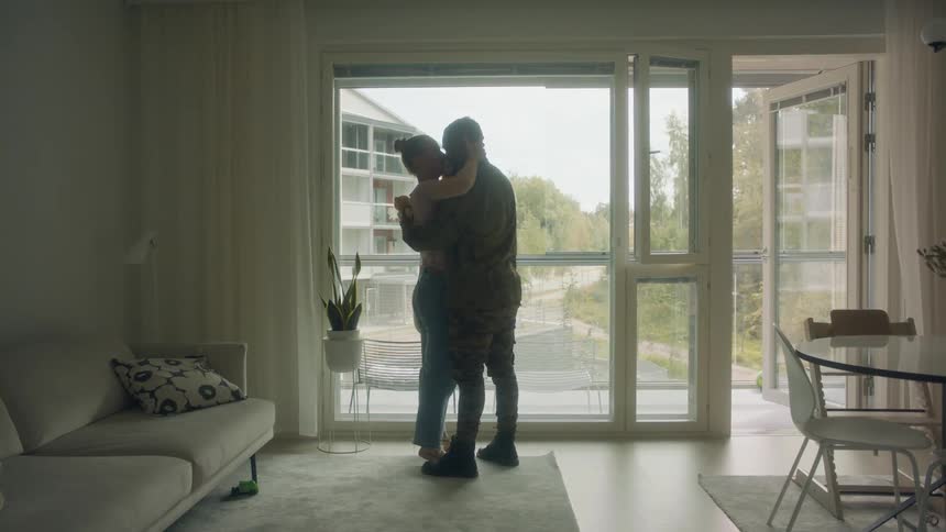 A soldier embraces and kisses a woman holding a baby while standing in the living room of a house