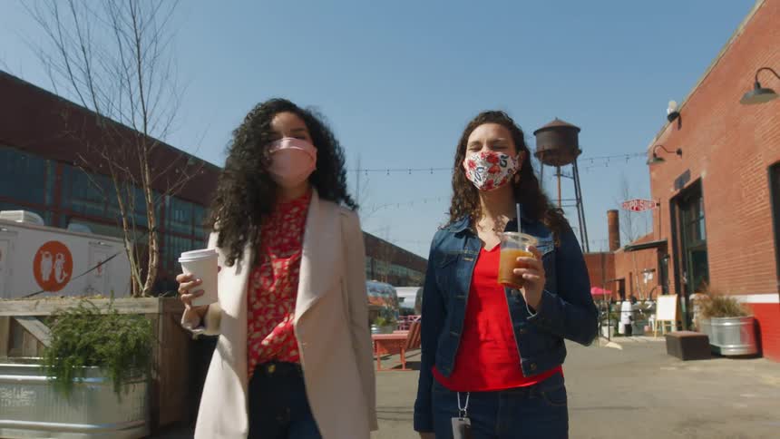 Two women are walking down a street together wearing masks and carrying drinking cups