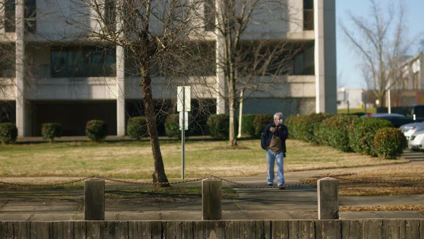 A man with a mask and a backpack walking with a building trees the sky and clouds in the background